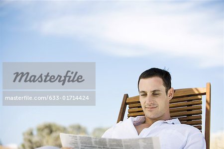 Young man sitting in deck chair, reading newspaper