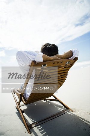 Man sitting in chair on beach hands, hands behind head, rear view