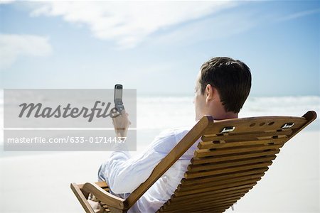 Man sitting in chair on beach, using cell phone, rear view