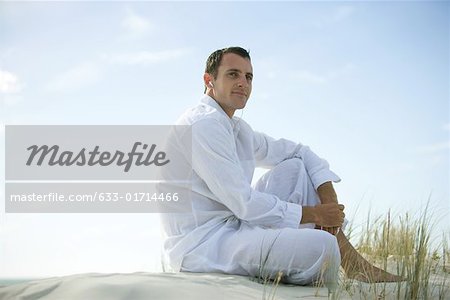 Young man sitting on dune, listening to earphones, smiling, looking away