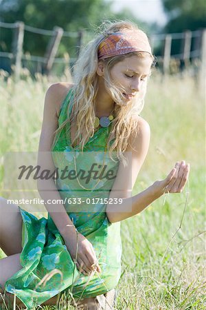 Young woman in sundress crouching in field, looking at sprig of grass