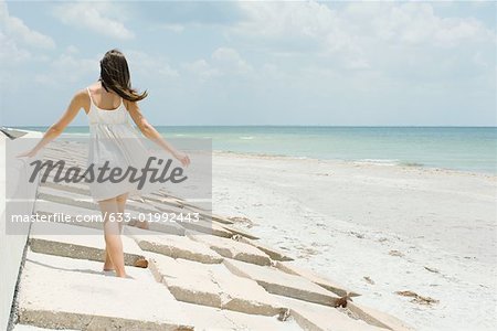 Young woman walking along low wall at the beach, rear view