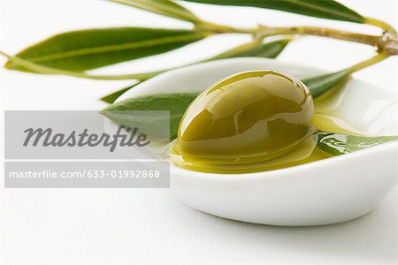 Green olive and sprig of leaves in small dish of olive oil, close-up