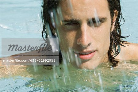 Man in swimming pool, looking at camera through splashing water, close-up