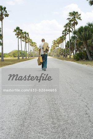 Man walking in center of road, carrying suitcase and jacket, rear view