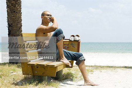 Man at the beach, sitting on bench beside suitcase, looking away