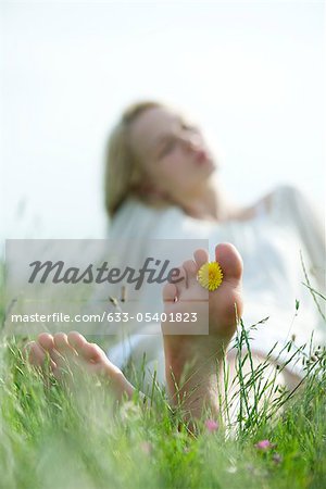 Barefoot young woman sitting in grass, holding dandelion flower between toes