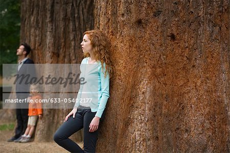 Mother leaning against tree trunk, husband and daughter separate in background