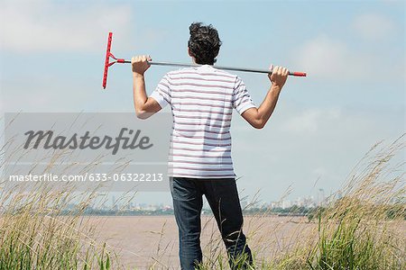 Man holding rake on shoulders, looking at city skyline from afar, rear view
