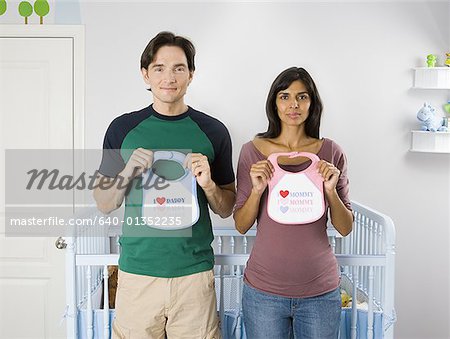 Portrait of a couple standing in front of a crib and holding baby bibs