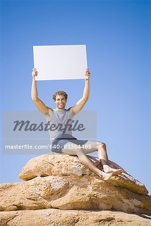Low angle view of a young man sitting on a rock with a blank sign holding up