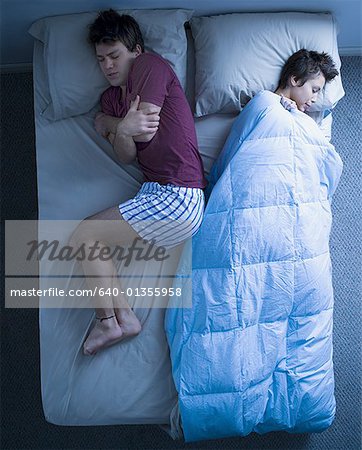 Woman in bed asleep with blanket and man sleeping curled up and cold
