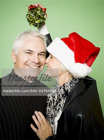 A woman holds mistletoe over a man's head while kissing him on the cheek