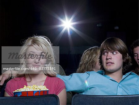 Boy reaching arm out behind girl with popcorn at movie theater