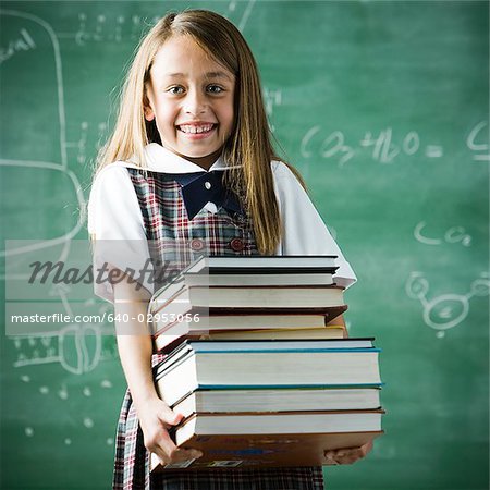 girl in a classroom standing in front of a chalkboard holding a stack of books