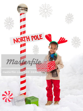 Boy (4-5) wearing reindeer antlers standing next to North Pole sign