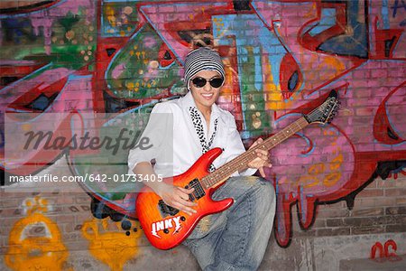 Young man wearing sunglasses playing guitar in front of a wall with graffiti