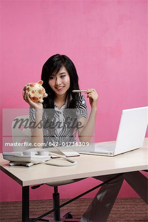 Young woman sitting by table and holding piggy bank, smiling