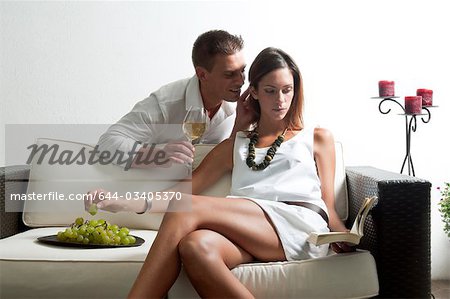 Young man leaning on couch with white wine whispering in young woman's ear reading book and eating grapes