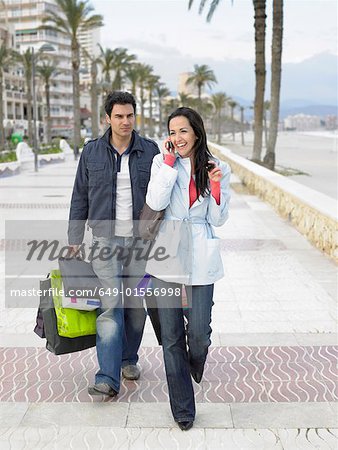 Couple walking along palm lined pavement, man carrying shopping bags while woman talks on mobile phone. Alicante, Spain.