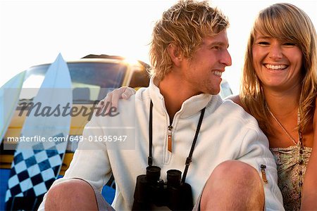 Couple sitting on beach smiling with van and surfboards in background.