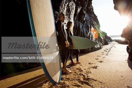 Couple standing with surfboards by large rocks.