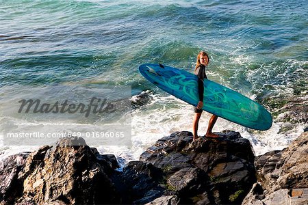 Woman standing on large rocks with a surfboard smiling.