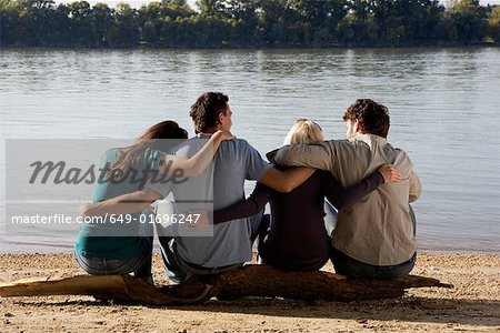 Four friends sitting on log by a lake with arms around each other.