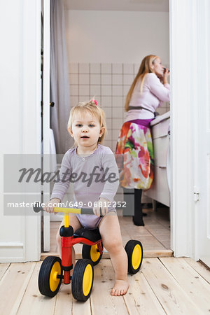 Toddler girl playing on tricycle, mother in background