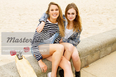 Girlfriends sitting on wall at beach with skateboard