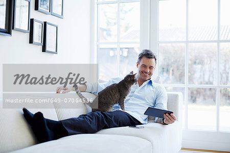 Man relaxing on sofa using digital tablet with cat