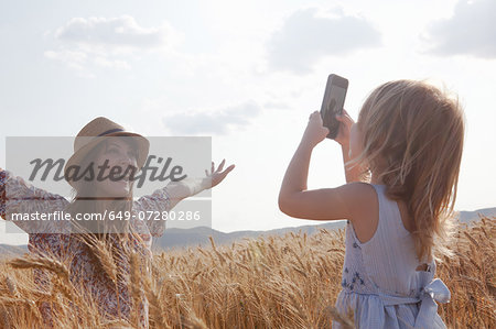 Girl taking photograph of mother in wheat field with arms open Girl taking photograph of mother in wheat field with arms open