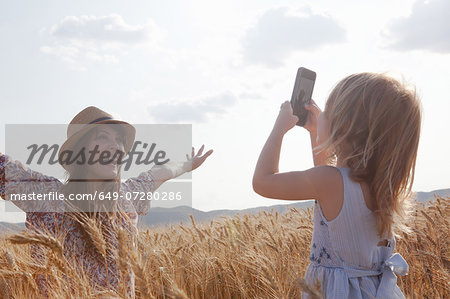 Girl taking photograph of mother in wheat field with arms open