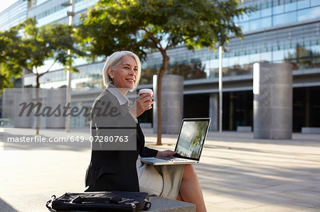 Businesswoman sitting outside using laptop and drinking hot drink