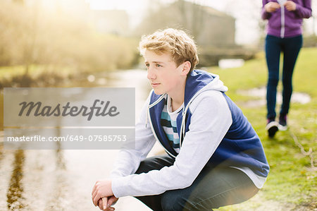 Unhappy teenage boy crouching on riverbank
