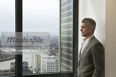 Portrait of mature businessman looking out of office window, Canary Wharf, London, UK