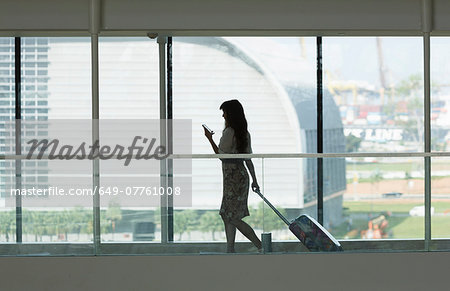 Young woman using smartphone and pulling suitcase in airport