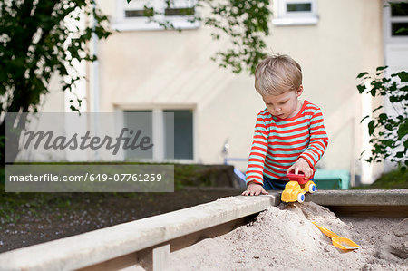 Male toddler pushing toy car in sand pit in garden