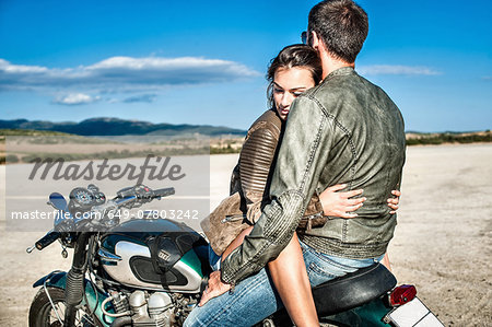 Young couple straddling motorcycle on arid plain, Cagliari, Sardinia, Italy