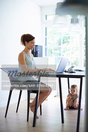 Mid adult mother typing on laptop with toddler daughter under the table Mid adult mother typing on laptop with toddler daughter under the table