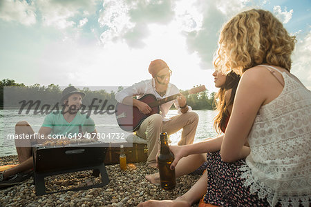 Young man sitting by lake with friends playing guitar