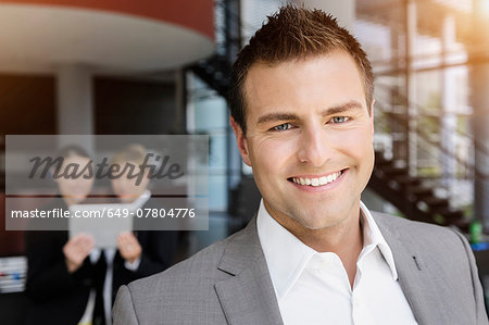 Smiling businessman, colleagues in background using digital tablet