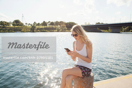 Young woman sitting at riverside texting on smartphone, Danube Island, Vienna, Austria