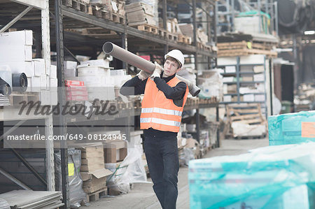 Young male warehouse worker carrying industrial pipe on shoulder