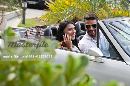 Mid adult couple in convertible car, woman using smartphone