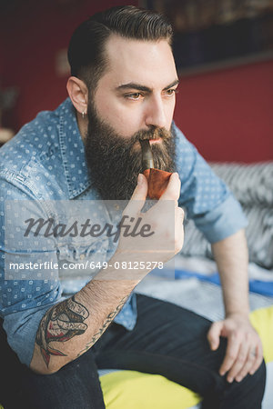 Young bearded man smoking pipe on bed