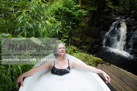 Mature woman relaxing in bubble bath in front of waterfall at eco retreat