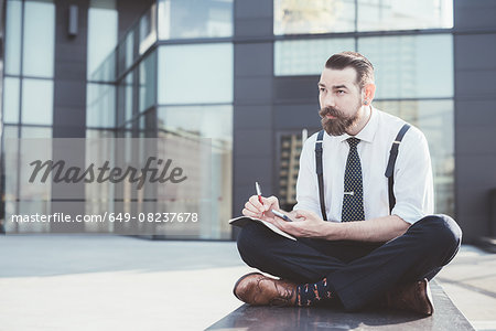 Stylish businessman sitting cross legged making diary notes from smartphone outside office