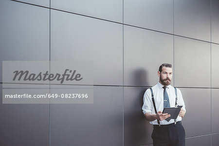 Stylish businessman using digital tablet and smartphone leaning against office wall