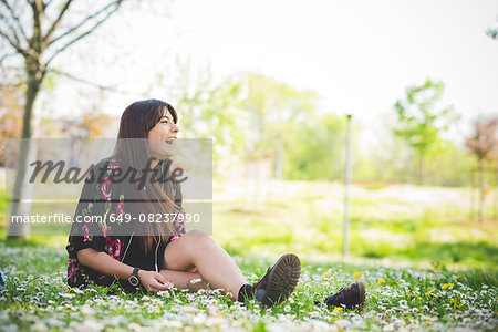 Young woman sitting on park grass listening to earphones